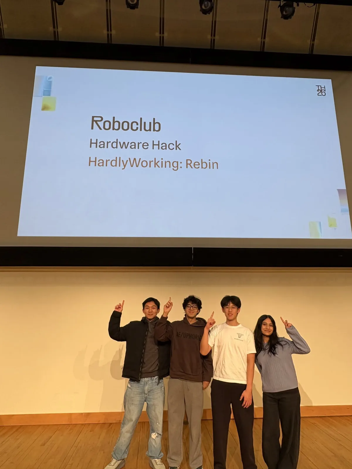 Four people on a stage under a projected slide reading Roboclub Hardware Hack, HardlyWorking: Rebin, each holding up one finger