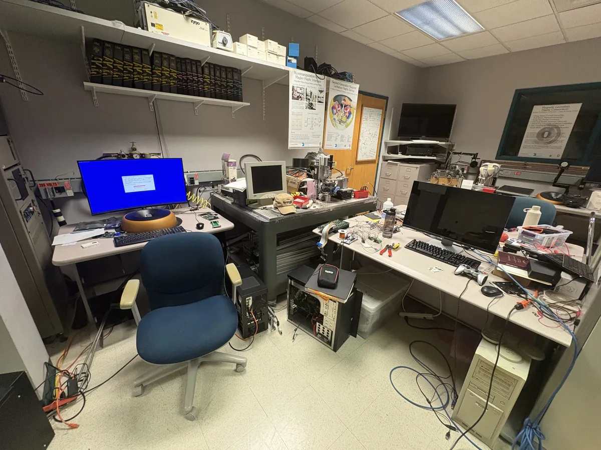 A lab room with two desks: on the left a monitor above an orange-and-black maglev haptic device and a keyboard, PC towers on the floor between the desks, and posters about maglev haptic interfaces on the wall