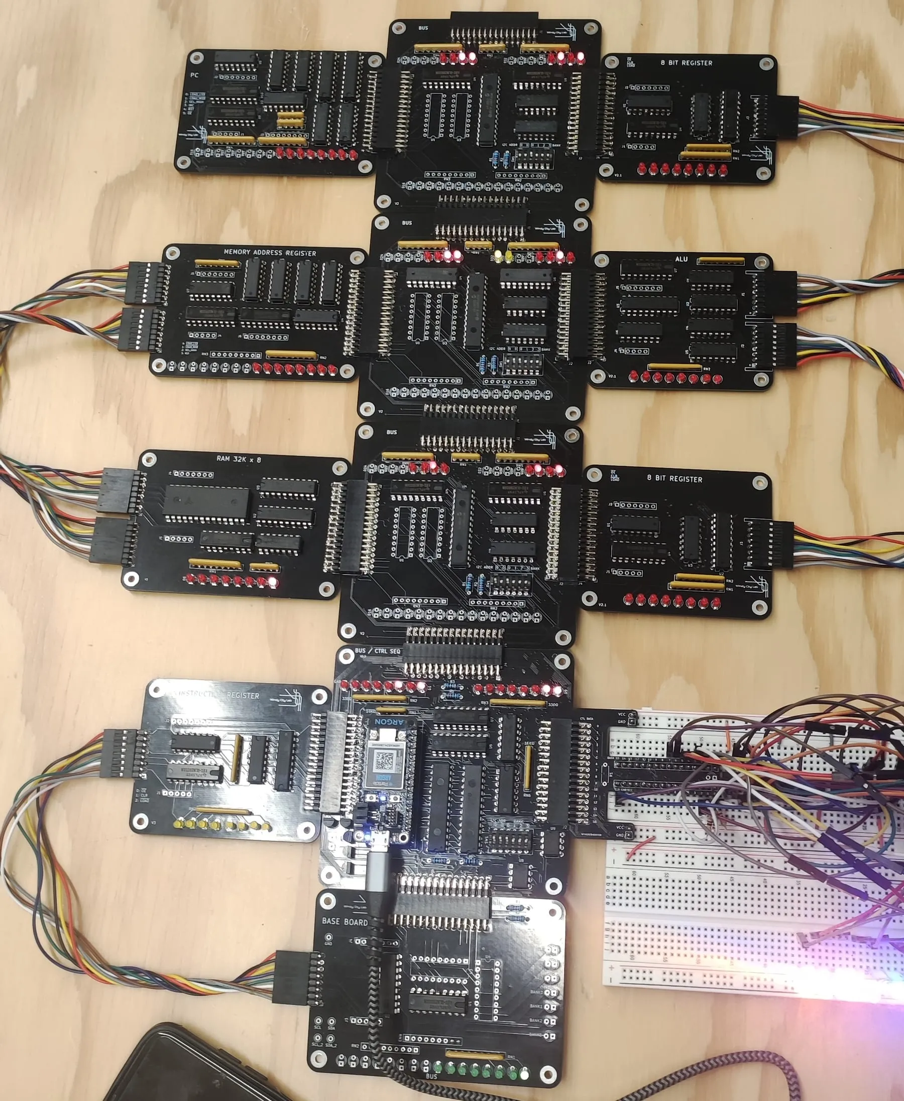 The 8-bit CPU laid out on a plywood bench as a grid of black PCBs: program counter, three bus boards, two 8-bit registers, memory address register, ALU, 32K×8 RAM, instruction register, bus/control sequencer and base board, with red status LEDs lit and ribbon cables running to a breadboard