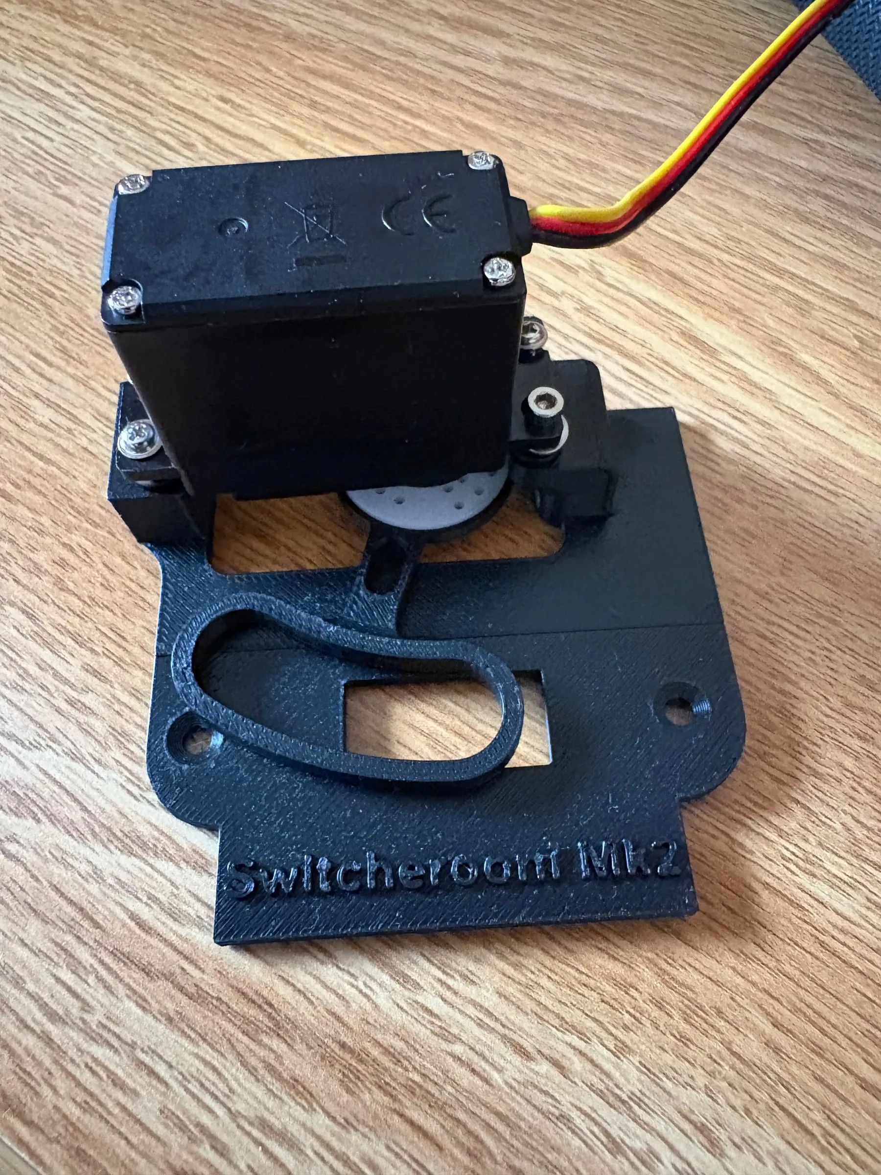 Switcherooni Mk2: a black 3D-printed plate embossed “Switcherooni Mk2”, with a servo mounted above the cutout for the light-switch toggle, on a wooden desk