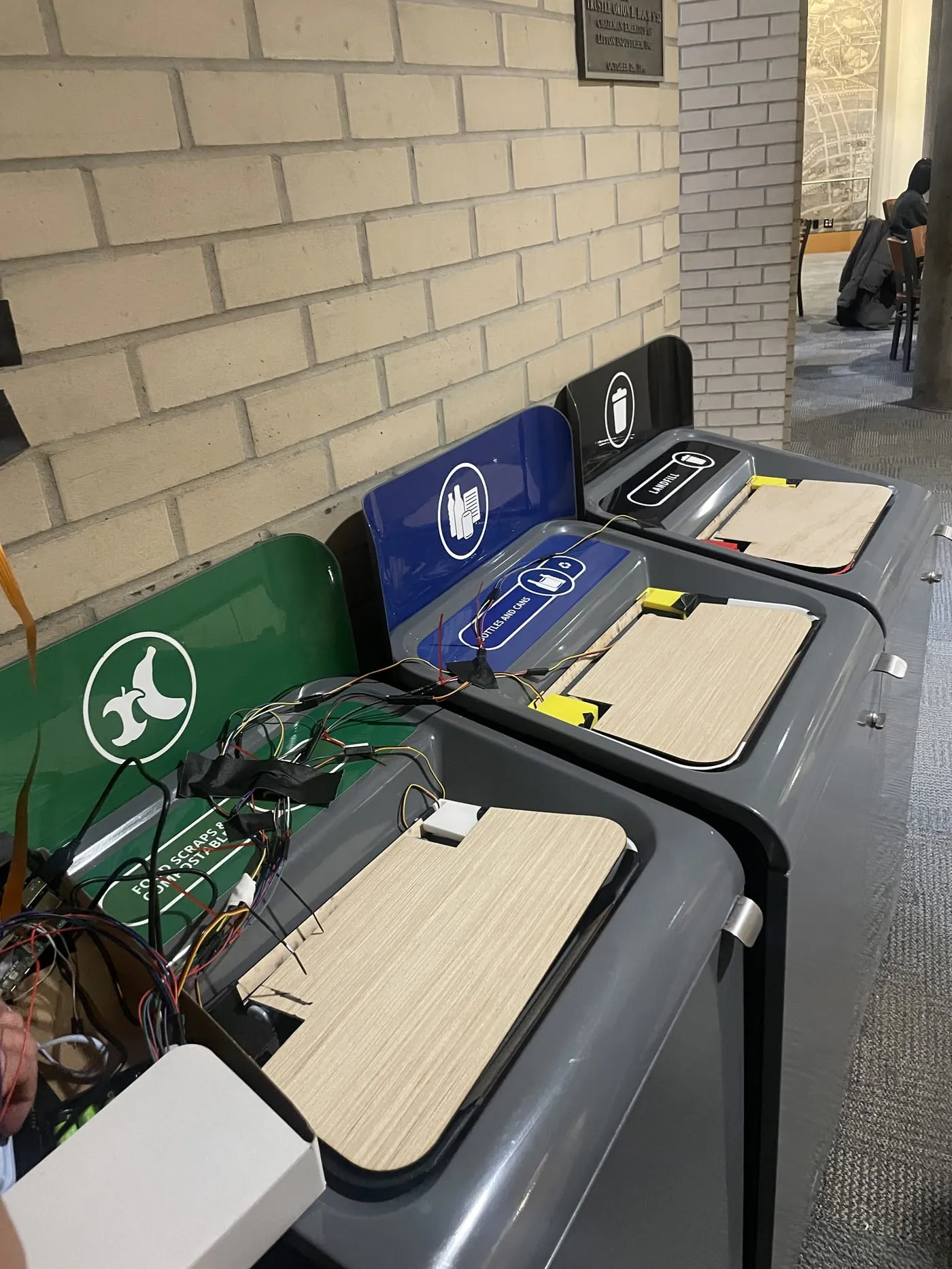 Three campus waste bins for compost, recycling and landfill, each fitted with a plywood lid and a bundle of wires taped along the top