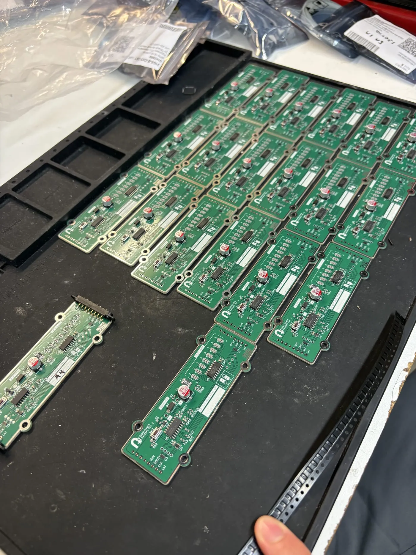 About twenty green RoboOrchestra handbell driver boards, Rev. 2, laid out in rows on a black tray beside bags of components