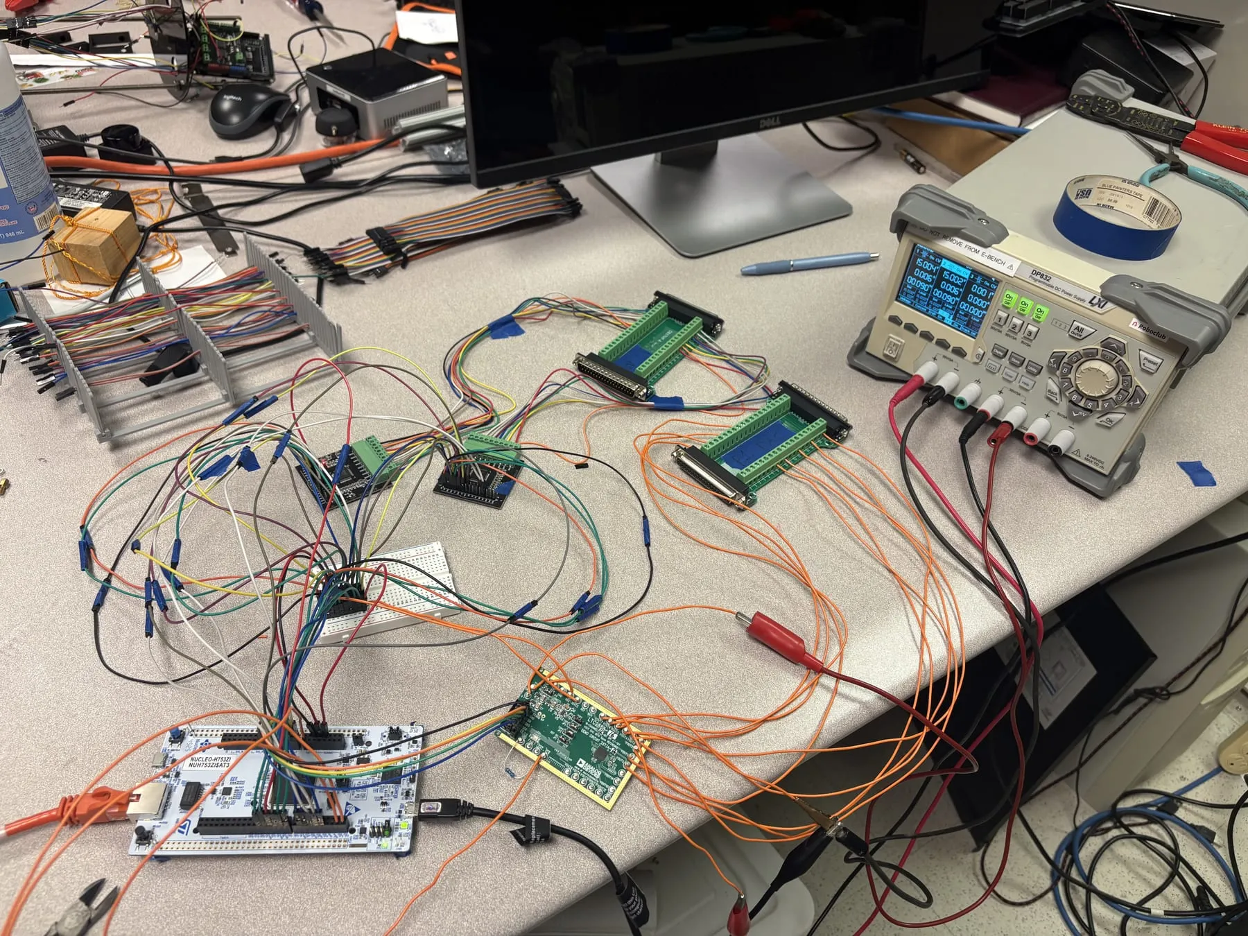 A lab bench: an STM32 Nucleo-H753ZI board wired with jumper leads to a breadboard, two DB37 breakout boards and an Analog Devices evaluation board, with a Rigol bench power supply behind them