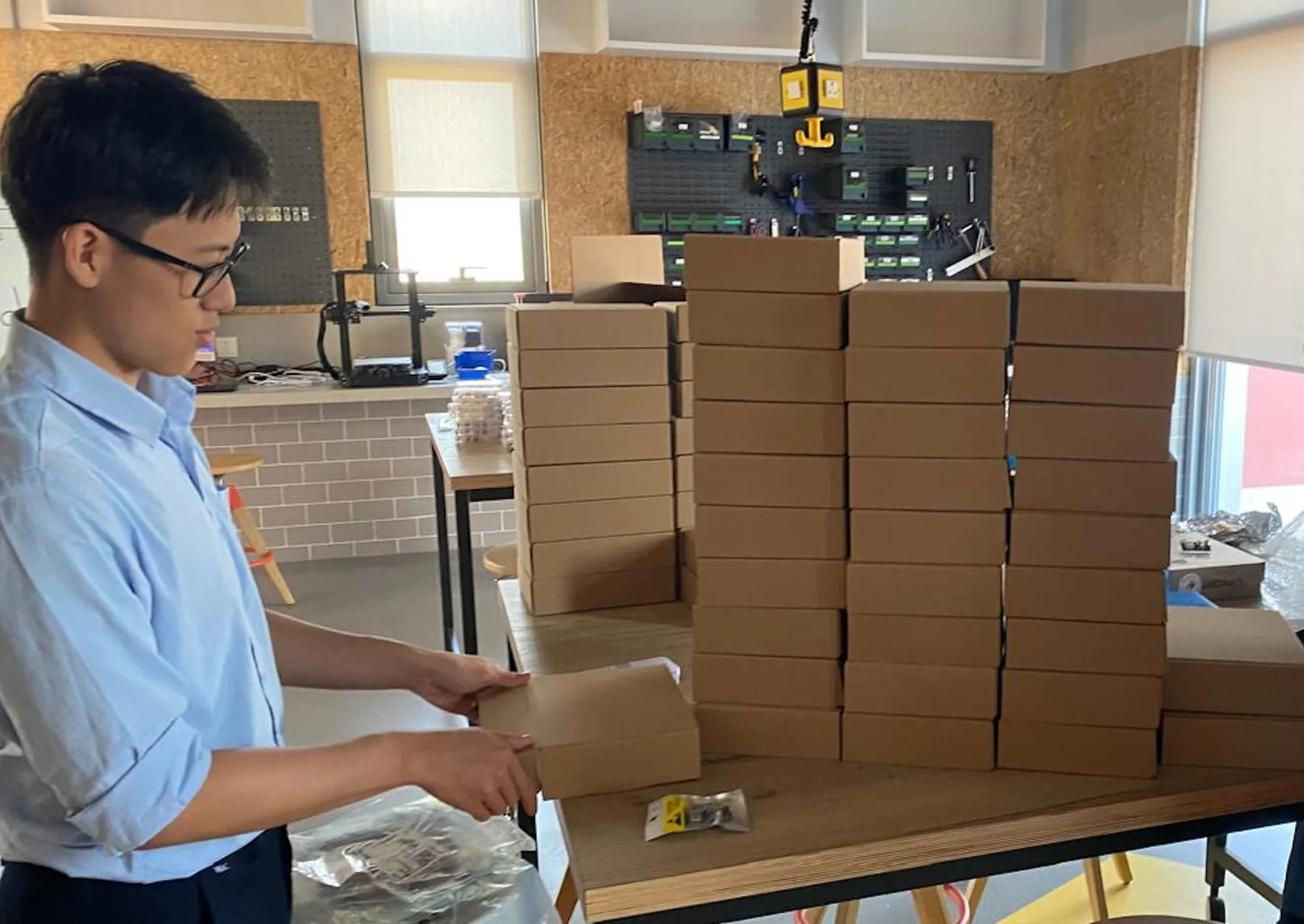 Zhaojin packing Logic Labs kits in the school makerspace: stacks of cardboard kit boxes on a workbench, a 3D printer and a component pegboard behind
