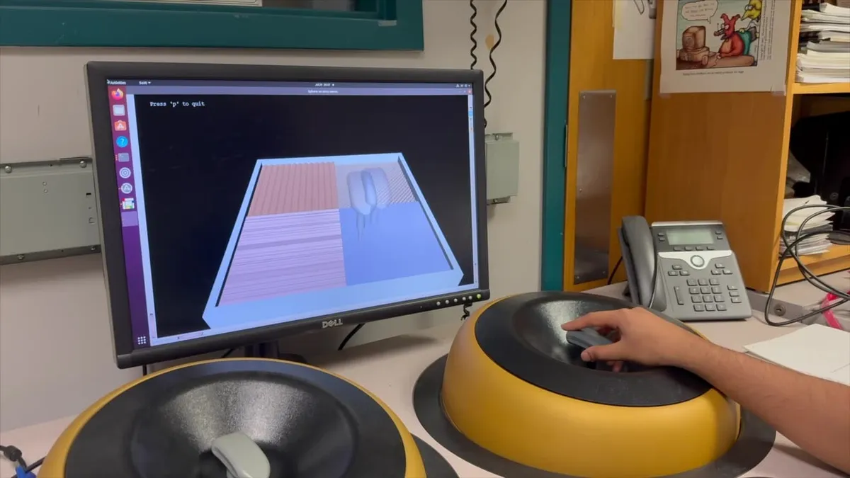 A monitor showing a SOFA simulation scene, a shaded box with a grey instrument inside it, beside a maglev haptic device with a hand resting on its handle