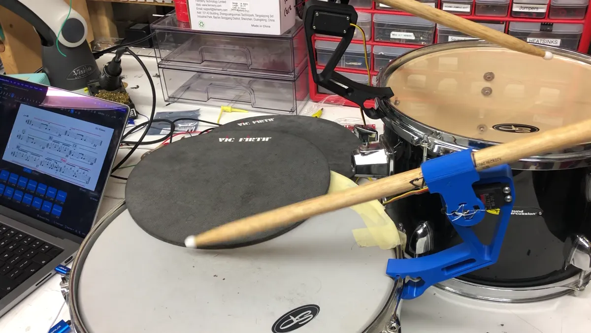 A drumstick held in a blue 3D-printed striker clamped to a snare drum, hitting a practice pad, with a laptop showing a score beside it