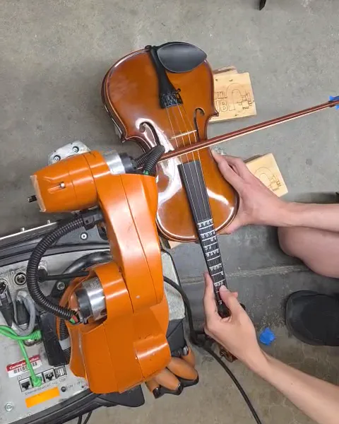 Seen from above, an orange robot arm drawing a bow across a violin held flat on a workbench